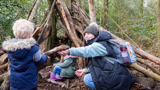 Oedolyn a phlentyn ger pentwr o bren sy'n creu siap cuddfan yn y goedwig
